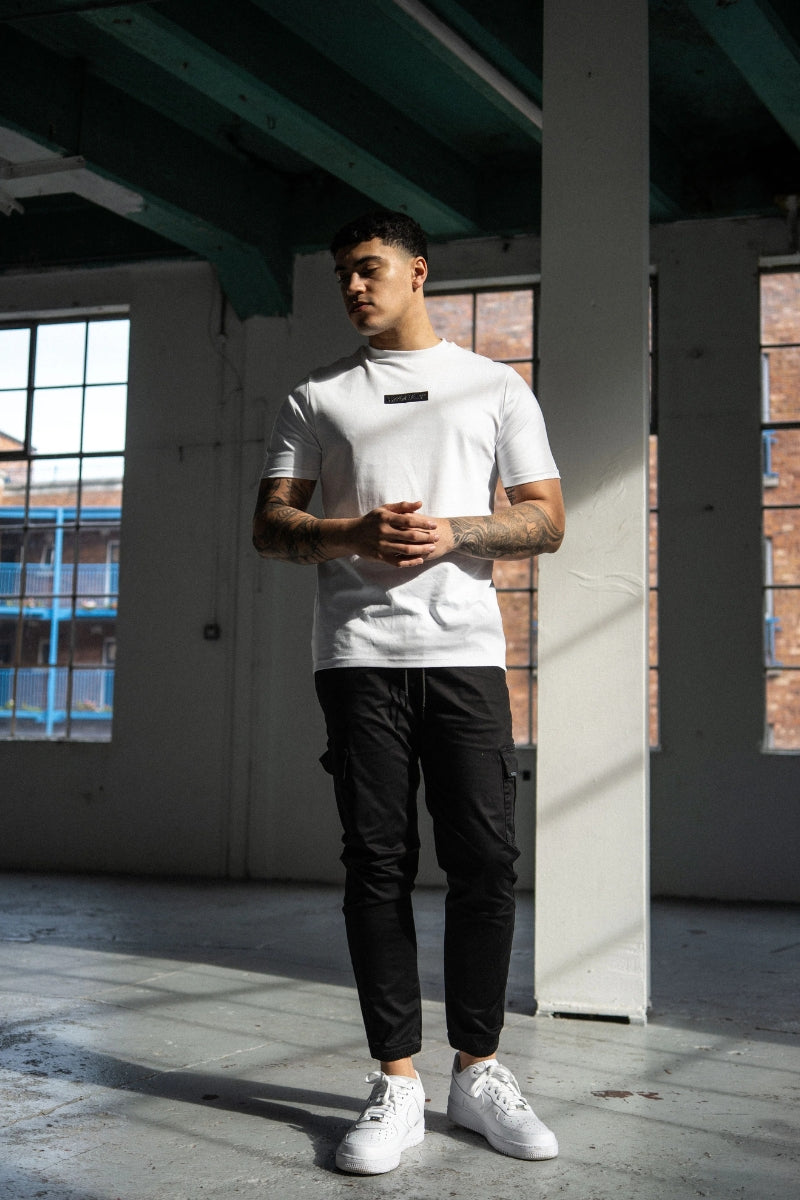 Man wearing a white t-shirt, black pants, and white sneakers in an industrial setting.