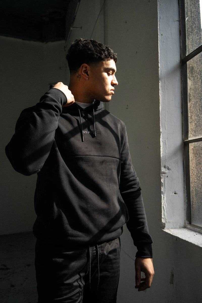 Man wearing a black hoodie standing in a dimly lit room with a window.