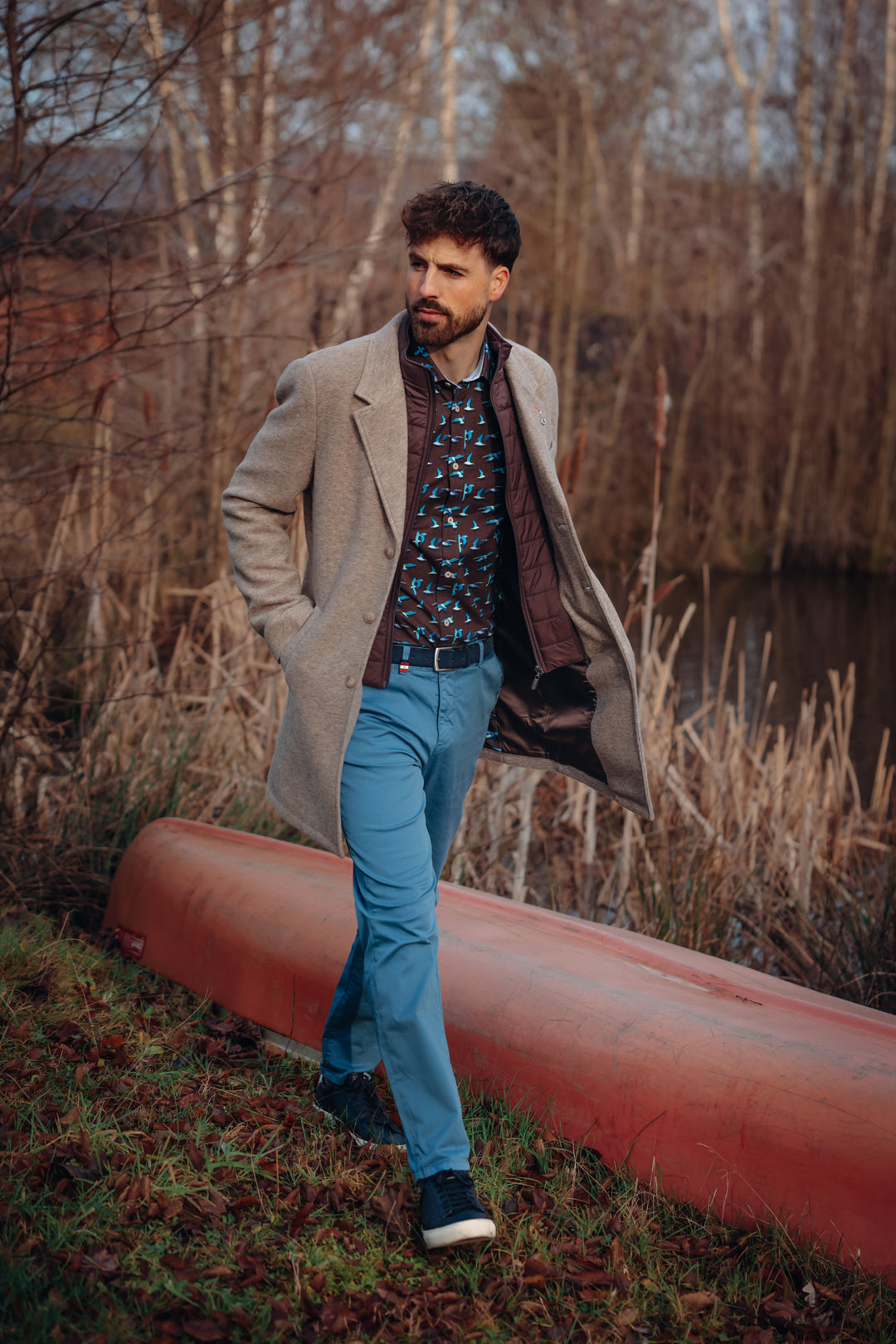 Man standing next to a red canoe in a natural setting wearing a sand colour coat