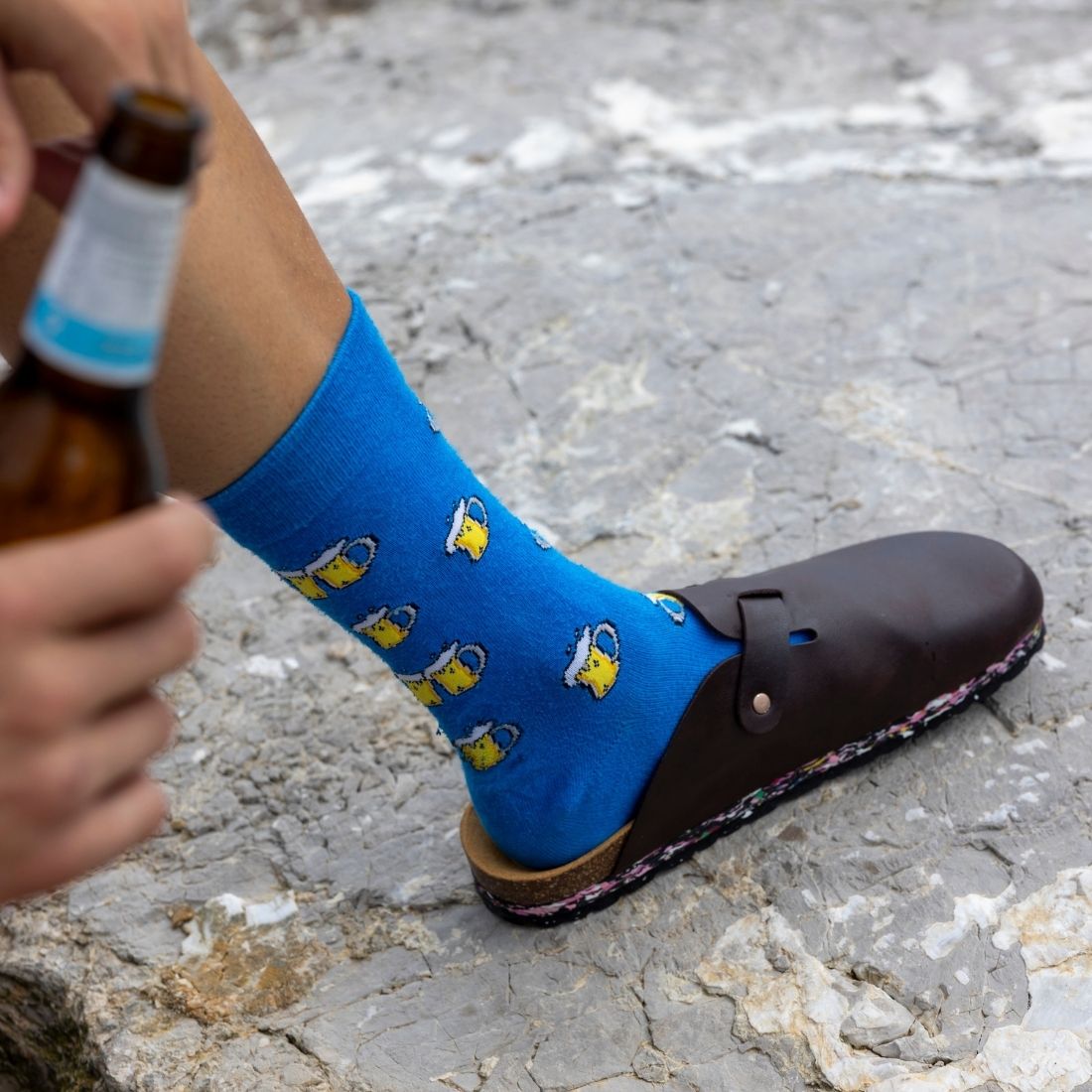 A person wearing a bright blue crew sock with a repeating pattern of yellow beer glasses and white foam, paired with a dark brown clog shoe, standing on a gray stone surface while holding a brown glass bottle.