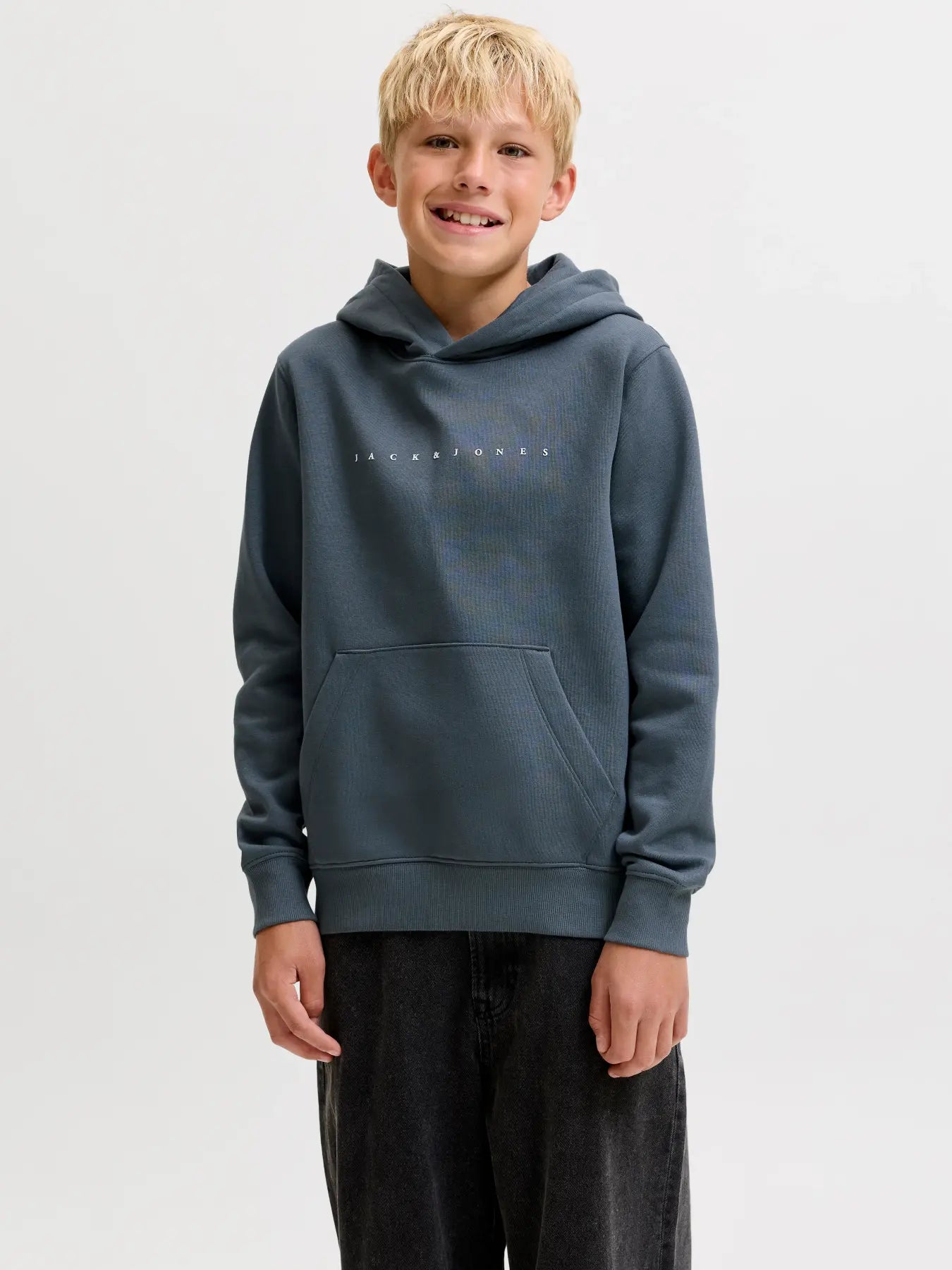 A young boy with blonde hair smiling, wearing a slate blue-gray pullover hoodie with a small white 'Jack Jones' logo across the chest, paired with dark gray jeans, against a light gray studio background.