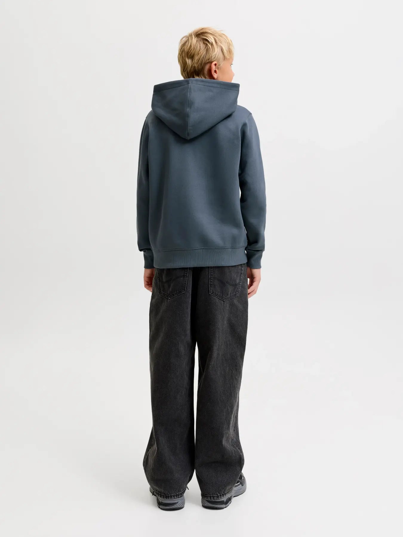 A child viewed from the back wearing a slate blue-gray pullover hoodie and dark wide-leg jeans against a plain light gray studio background.