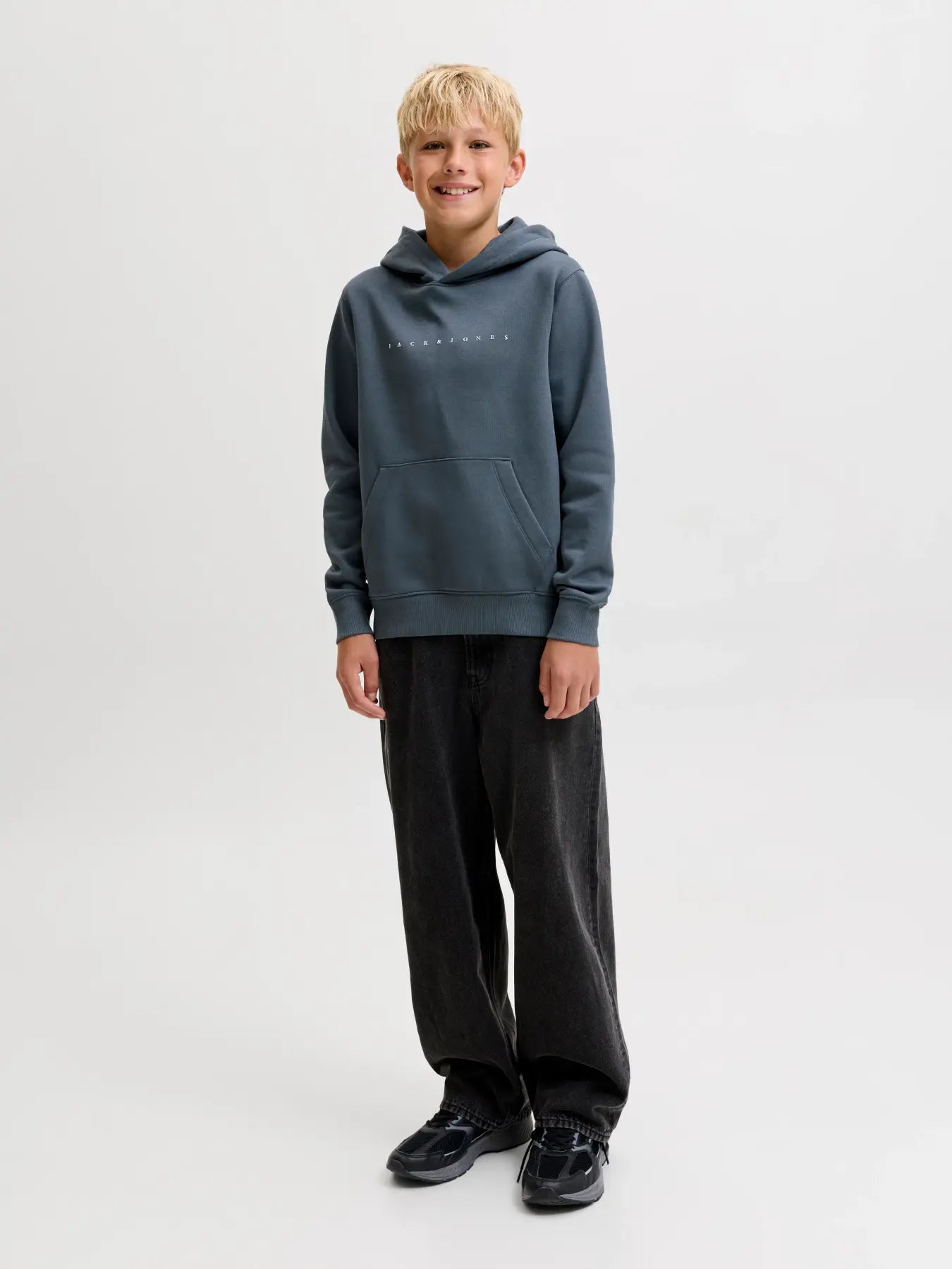 A young boy stands against a light gray studio background wearing a slate blue-gray pullover hoodie with a small white chest logo, black sweatpants, and black shoes.