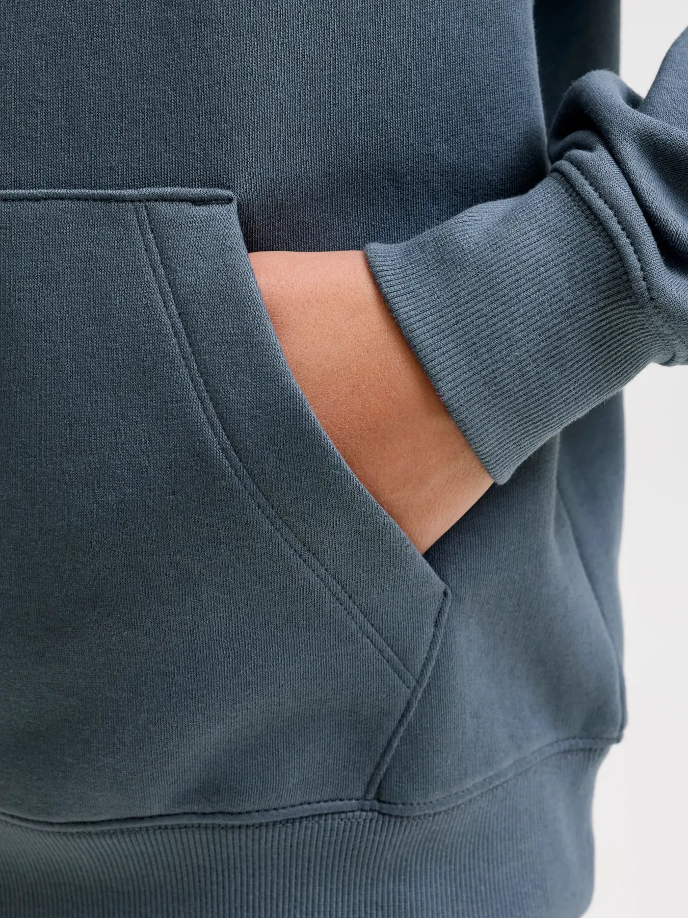 Close-up of a person wearing a slate blue-gray hoodie, showing the ribbed cuff of the sleeve with a hand tucked into a front kangaroo pocket.