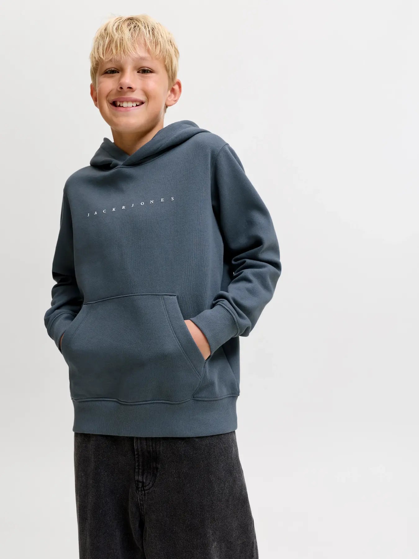A smiling young boy with blonde hair wearing a slate blue-gray pullover hoodie with a small white dotted logo on the chest, paired with dark gray jeans, against a plain light gray studio background.
