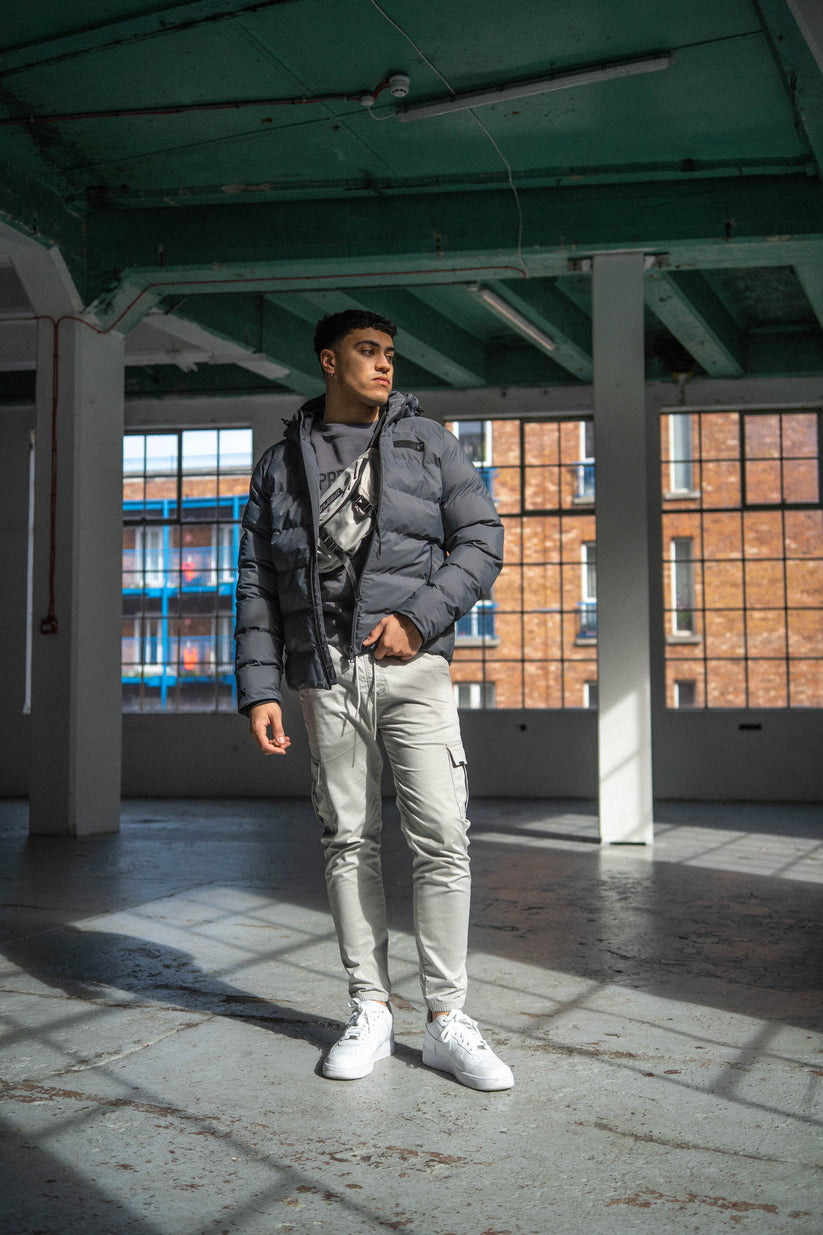 Man wearing a gray puffer jacket and light-colored pants standing in an industrial-style building.