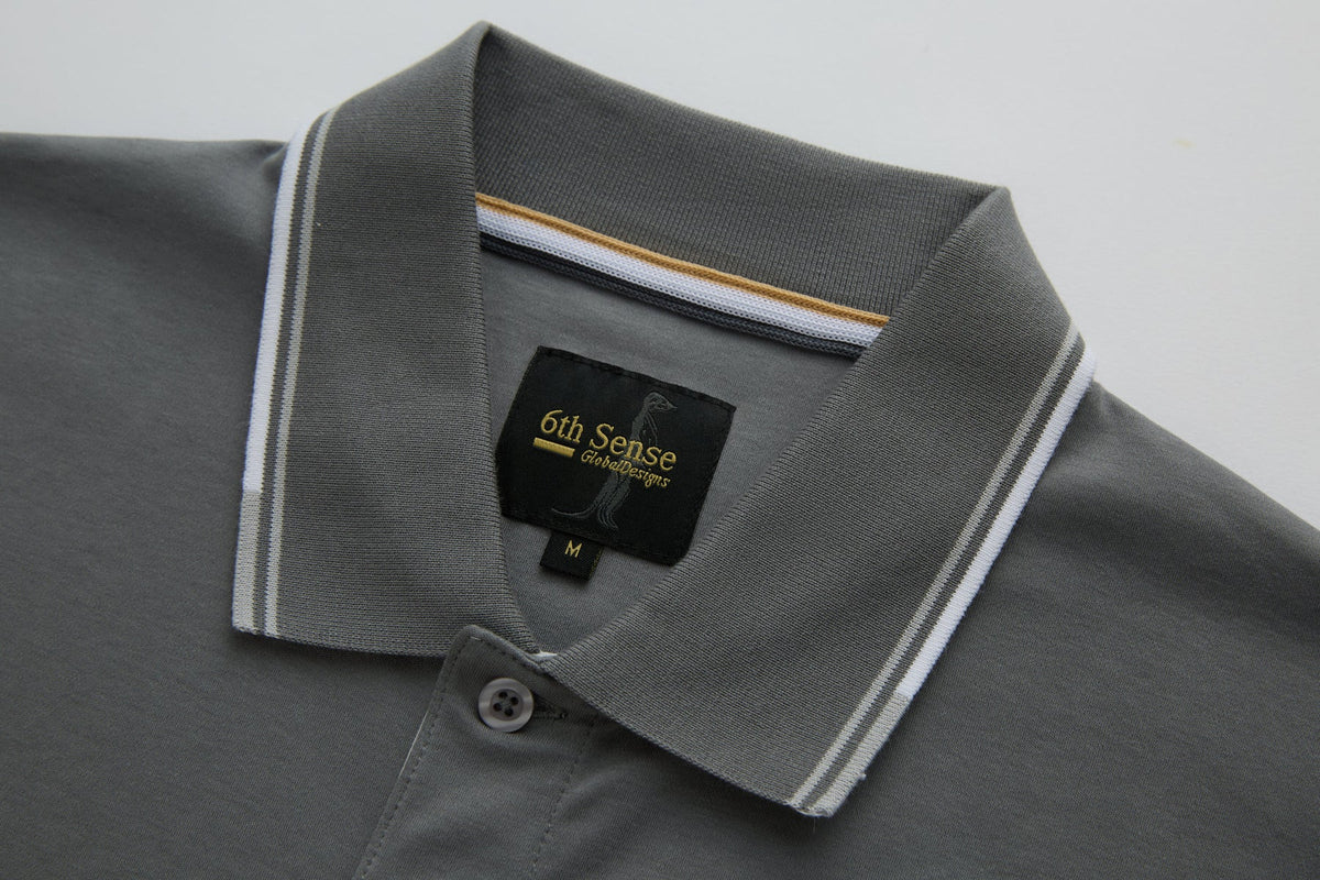 Close-up of the 6th Sense Astound December Polo in December, showing the collar, button placket, and inner neck label. The collar has white and brown striped trim, and the black woven label features the 6th Sense Global Designs logo with a size M tag below.
