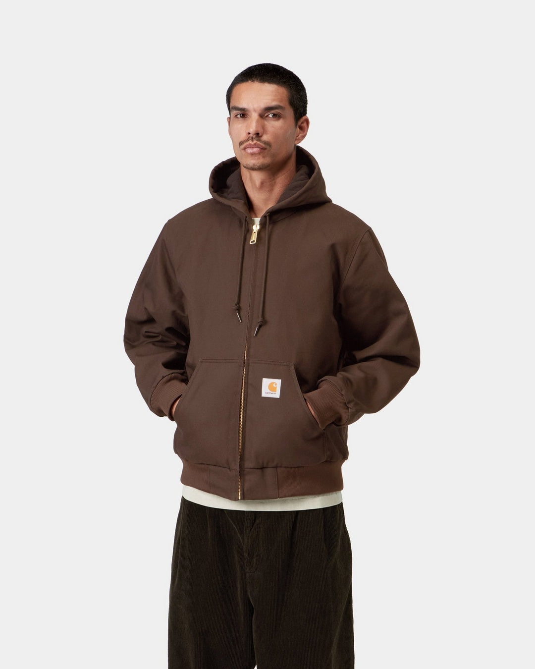 Front view of the Carhartt WIP Active Jacket in Tobacco, worn with the hood down and hands in the front pockets. The brown jacket features a gold-tone zip, drawstring hood, ribbed cuffs and hem, and a square Carhartt WIP logo patch on the kangaroo pocket.