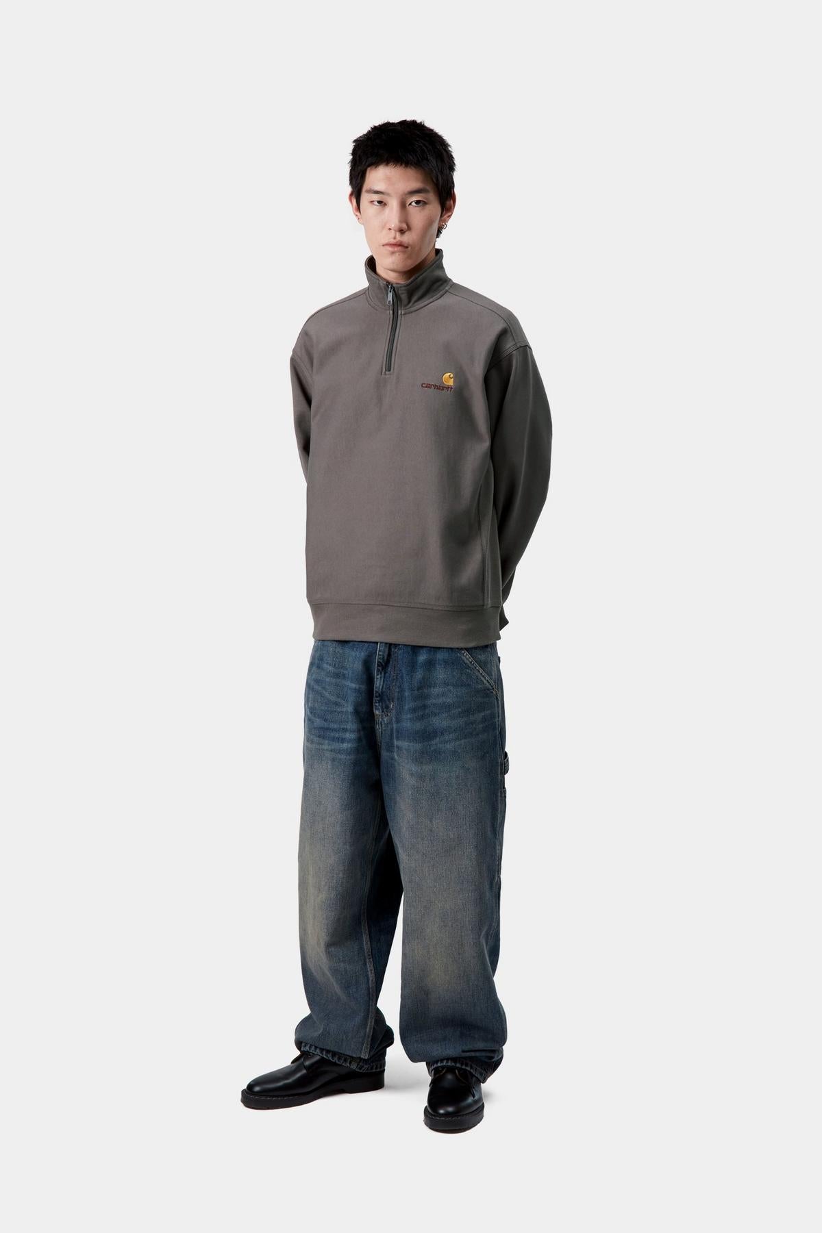 Person wearing a gray pullover with a logo and blue jeans on a white background