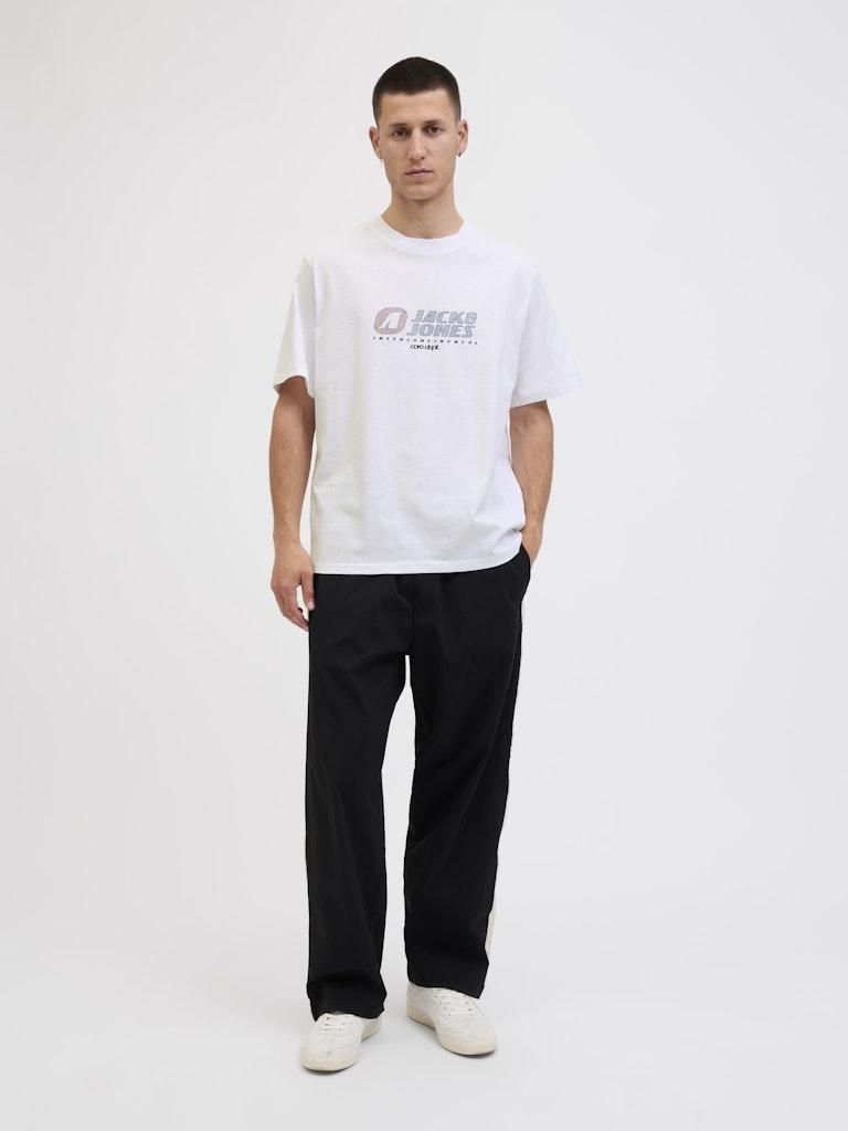 Man wearing a white t-shirt with a logo and text, black pants, and white shoes on a white background