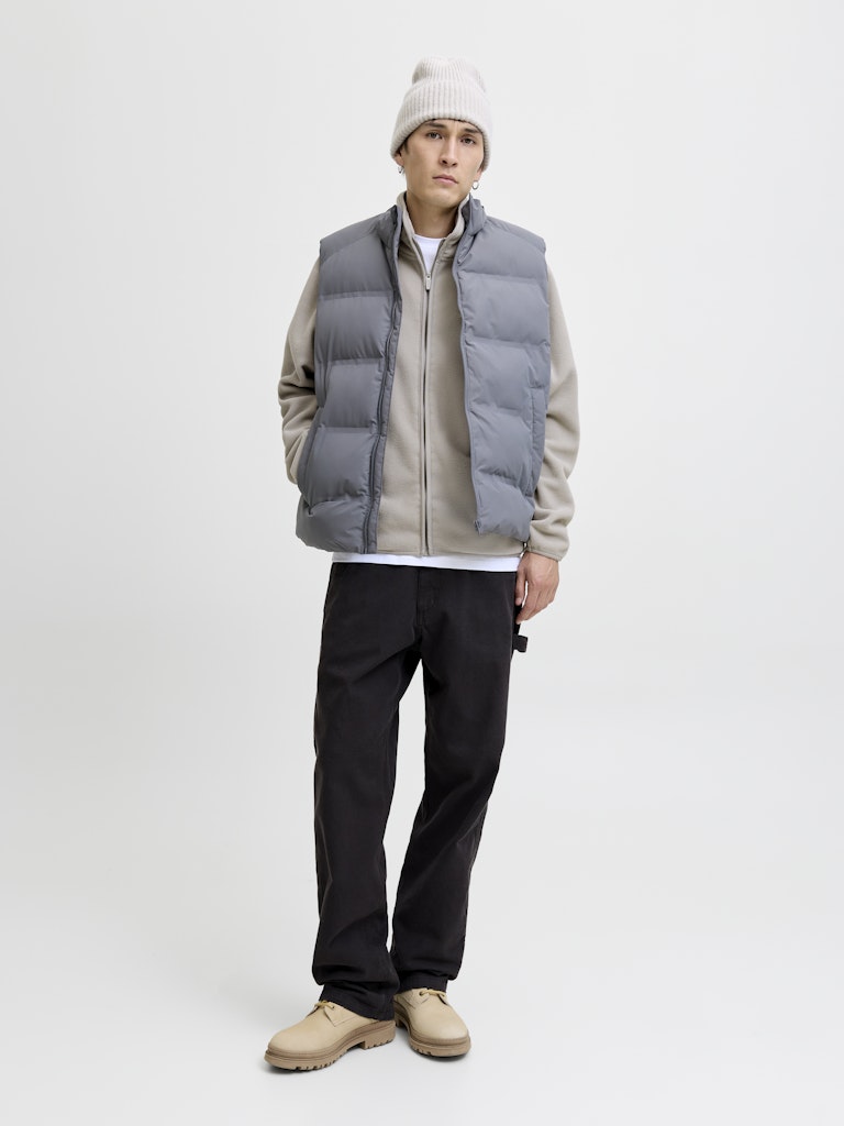 Person wearing a gray puffer vest over a beige shirt, black pants, and a white beanie on a white background