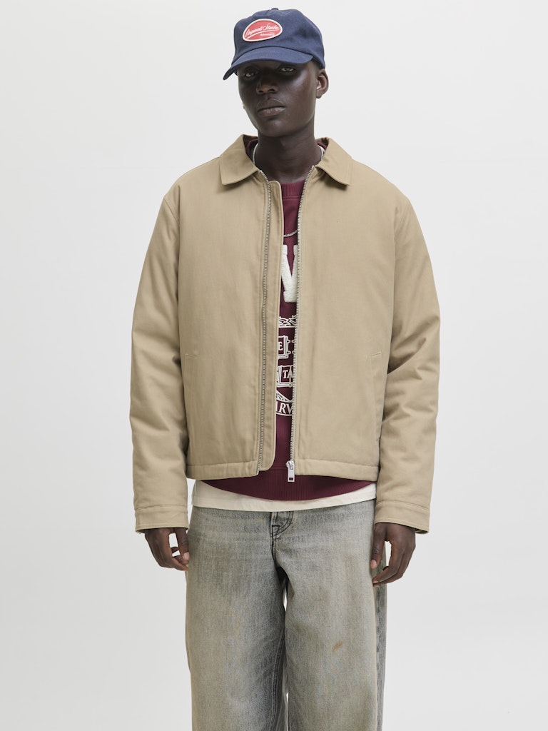 Man wearing a beige jacket, maroon shirt with text, and blue cap on a light gray background