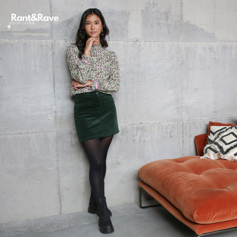 Woman in floral blouse and green skirt leaning against a textured wall with an orange couch and patterned pillow.