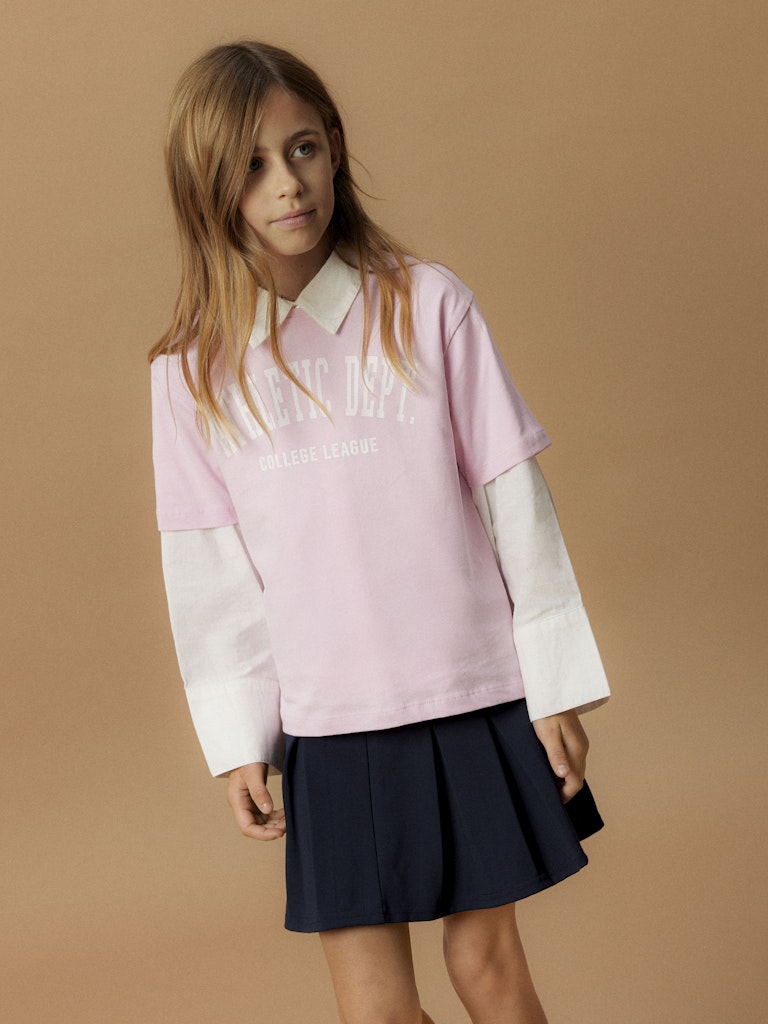 Person wearing a pink and white shirt with text and a navy pleated skirt on a beige background