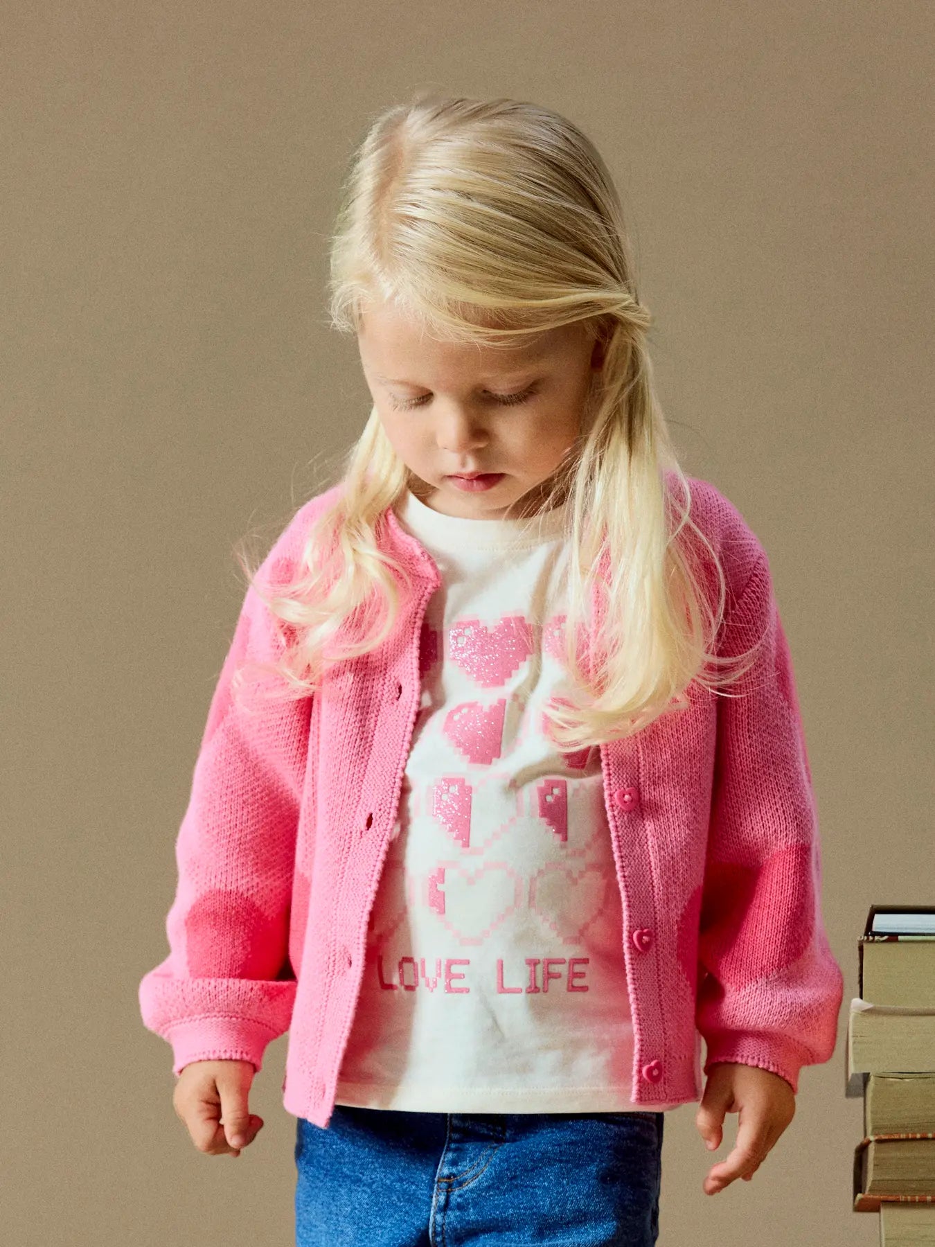 A young blonde girl wearing a pink knit cardigan over a white graphic t-shirt and blue jeans, standing against a solid beige background.