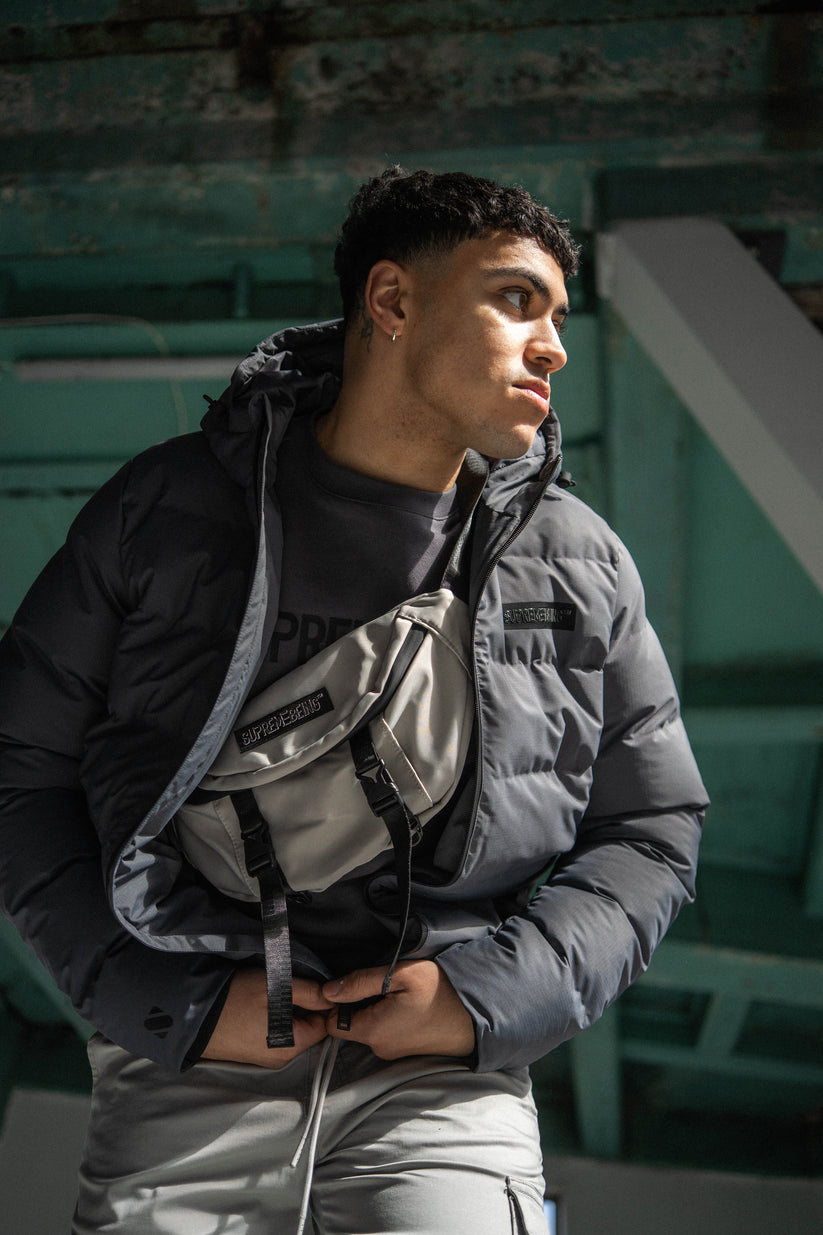 Person wearing a black and gray jacket with a bag, standing against a green wall.