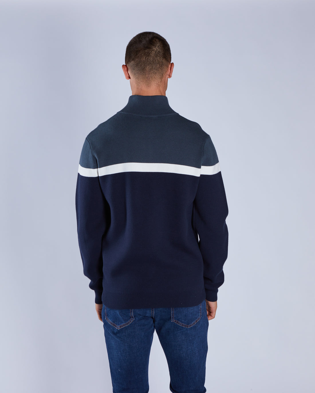 Man wearing a navy blue sweater with white stripes on the back against a light gray background