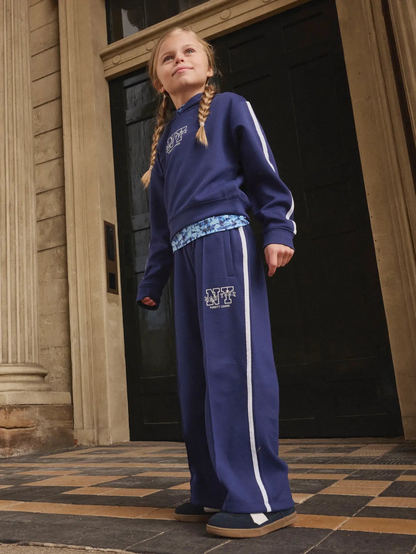 Young girl wearing a blue tracksuit with a logo, standing in front of a building entrance.