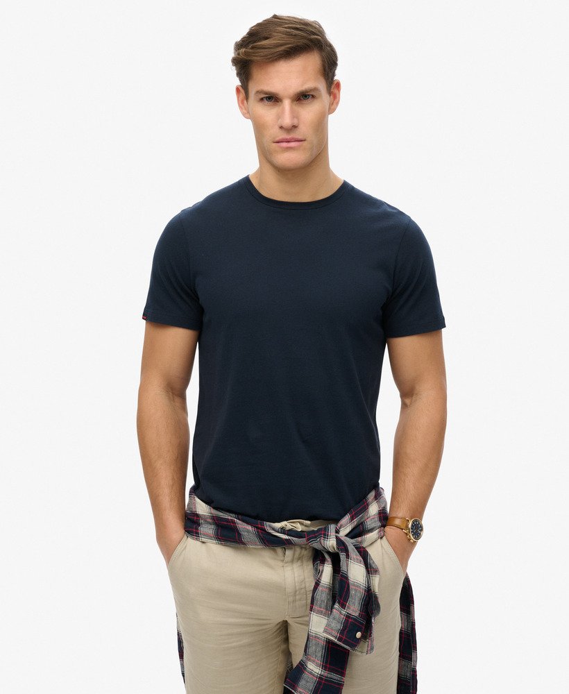 Man wearing a navy blue t-shirt and beige pants with a plaid shirt over his shoulder on a white background