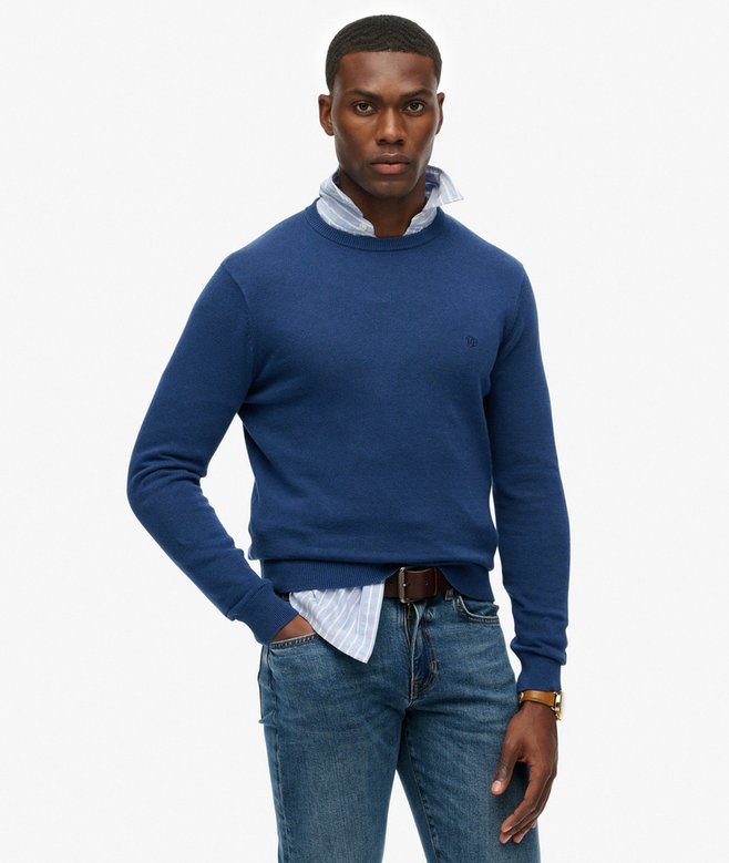 Man wearing a blue sweater over a white shirt with jeans on a white background