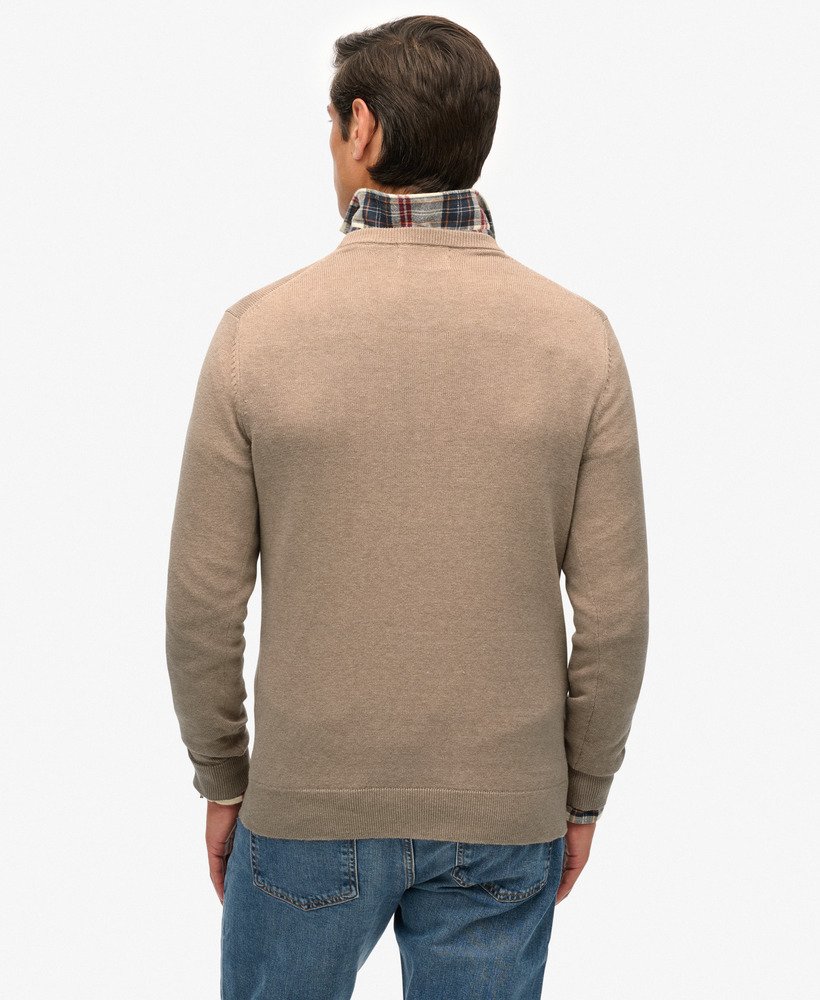 Man wearing a beige sweater over a plaid shirt with blue jeans on a white background