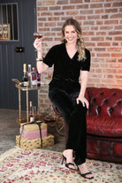 Rant & Rave Clara Trouser Black worn by seated model holding a drink, angled view, wide - leg velvet trousers with matching top