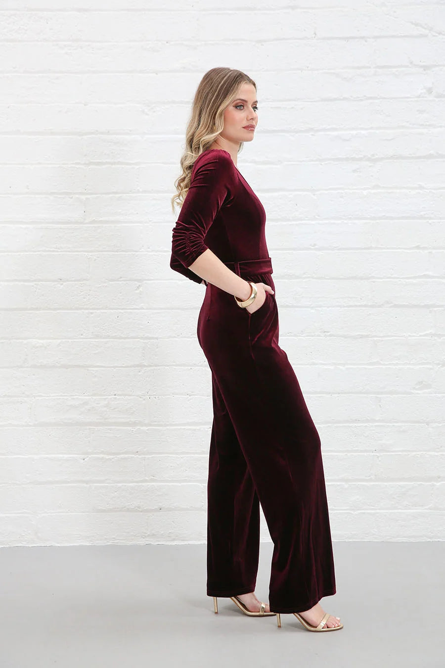 Rant & Rave Farrah Jumpsuit in wine velvet, side model view with hand in pocket and gold heels
