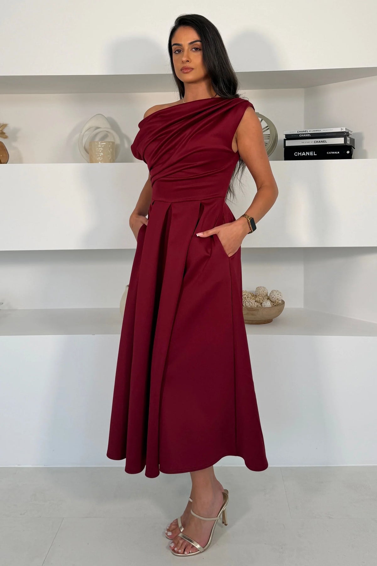 Girl In Mind Betty asymmetric neckline skater midi dress in wine, worn by model, side view with hands in pockets and pleated midi skirt
