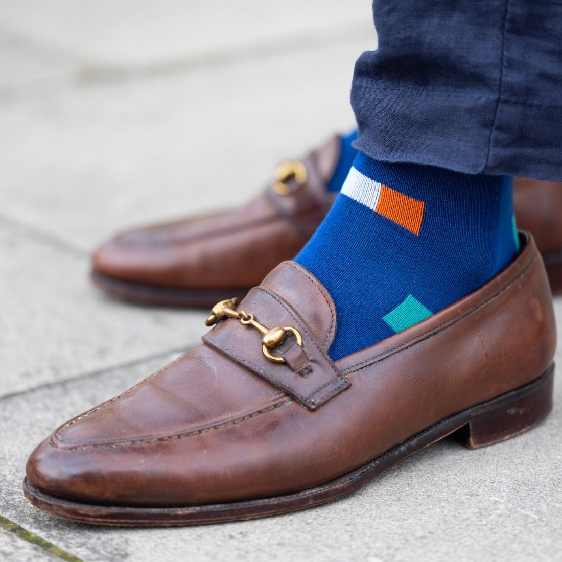 Brown loafers with blue socks featuring an Irish flag design on a concrete surface.