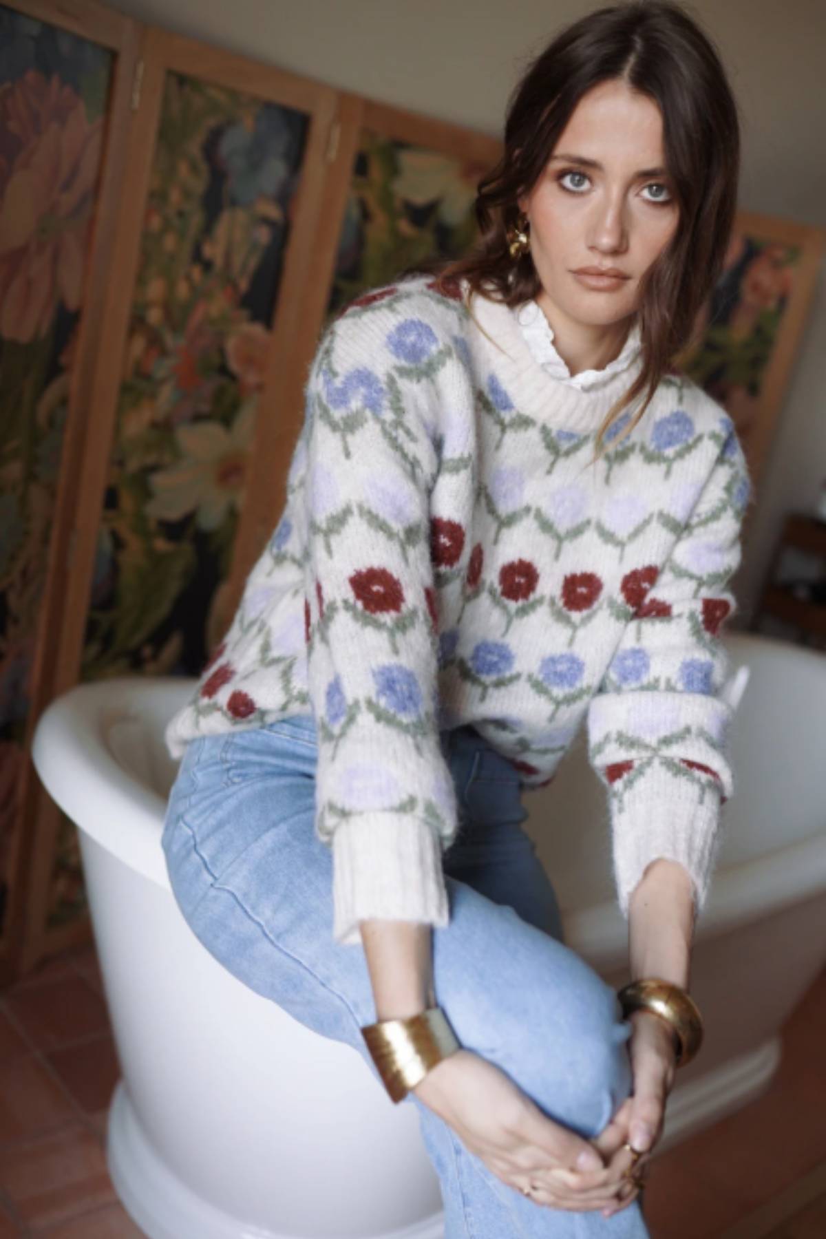 Woman sitting on a white bathtub wearing a floral sweater and blue jeans, with a colorful abstract painting in the background.
