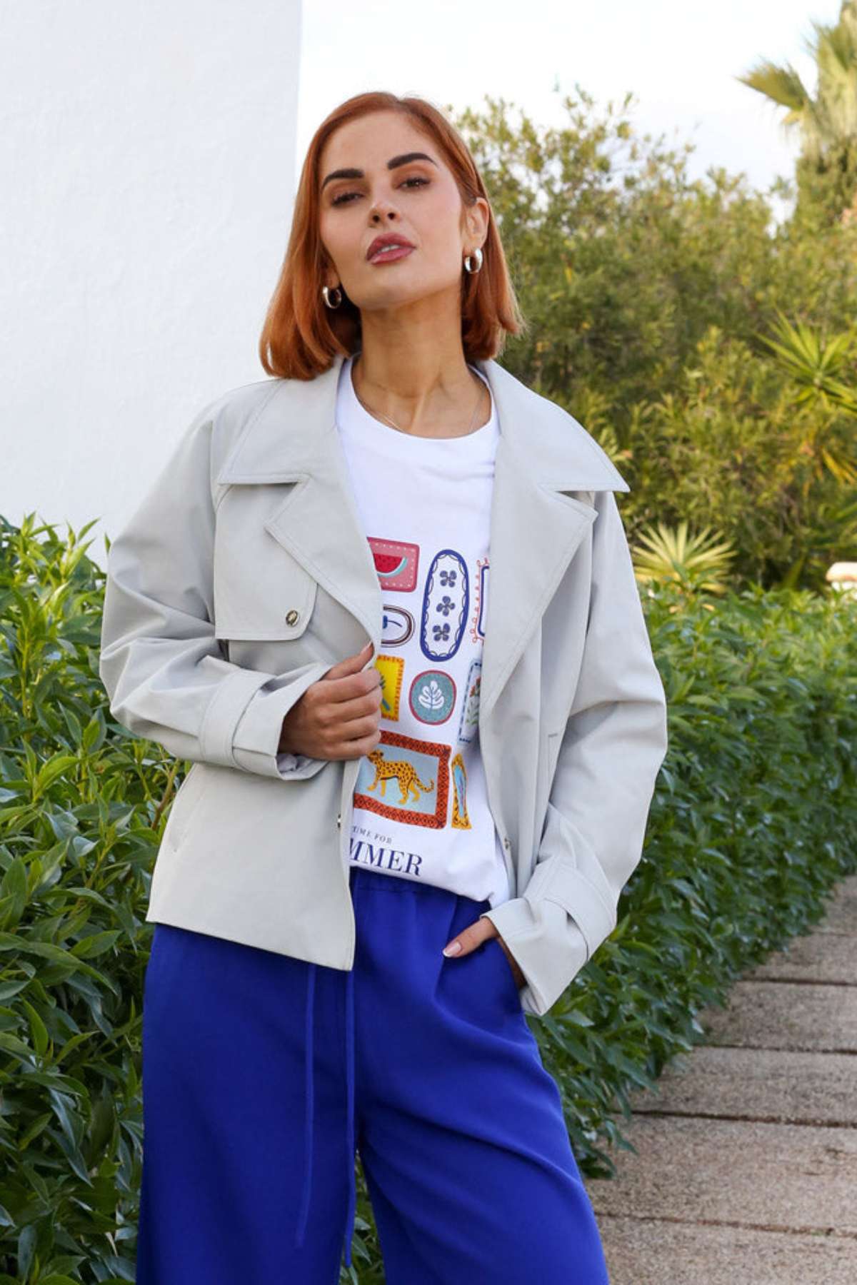 Woman wearing a light gray jacket over a colorful graphic t-shirt with blue pants, standing outdoors.