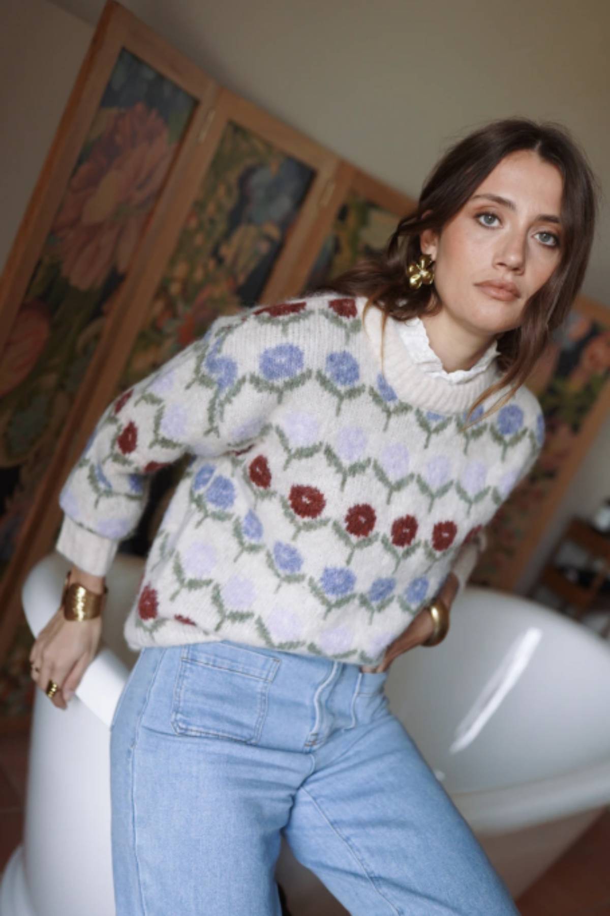 Woman wearing a patterned sweater and jeans in an indoor setting with artwork on the wall.