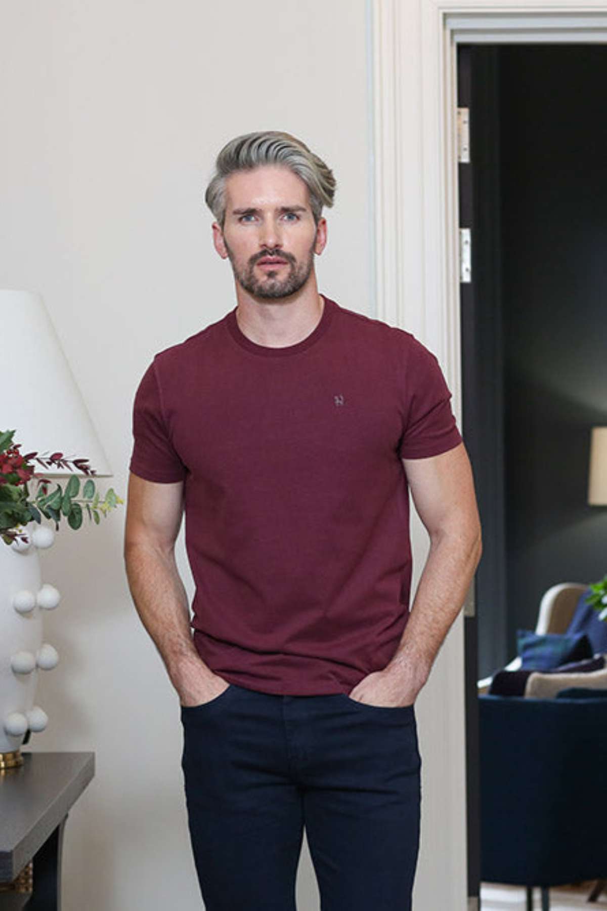 A man with styled gray hair and a beard wearing a maroon crewneck t-shirt and dark navy pants, standing in a modern indoor setting with a lamp on the left and a doorway in the background.