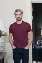 A man with styled gray hair and a beard wearing a maroon crewneck t-shirt and dark navy pants, standing in a modern indoor setting with a lamp on the left and a doorway in the background.
