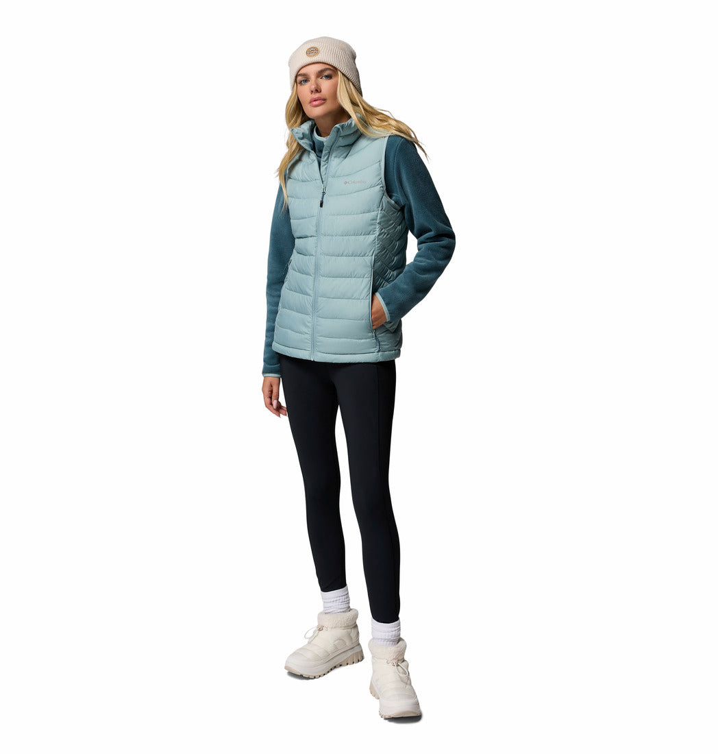 Woman wearing a teal puffer vest, black leggings, white shoes, and a beige beanie on a white background