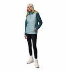 Woman wearing a teal puffer vest, black leggings, white shoes, and a beige beanie on a white background