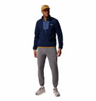 Man wearing a navy pullover, gray jogger pants, and a yellow cap on a white background
