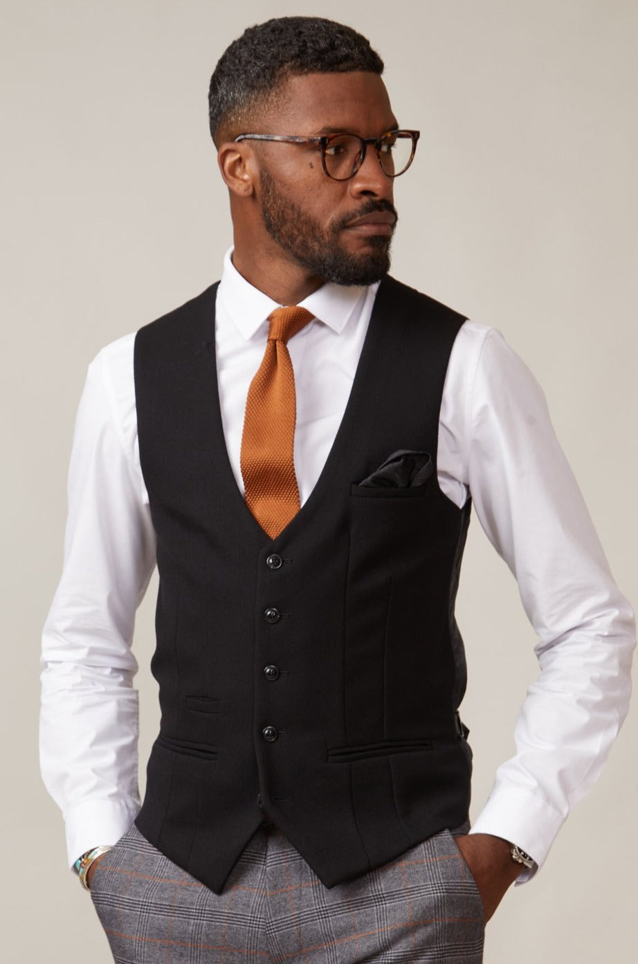 Man wearing a black waistcoat, white shirt, and brown tie against a plain background