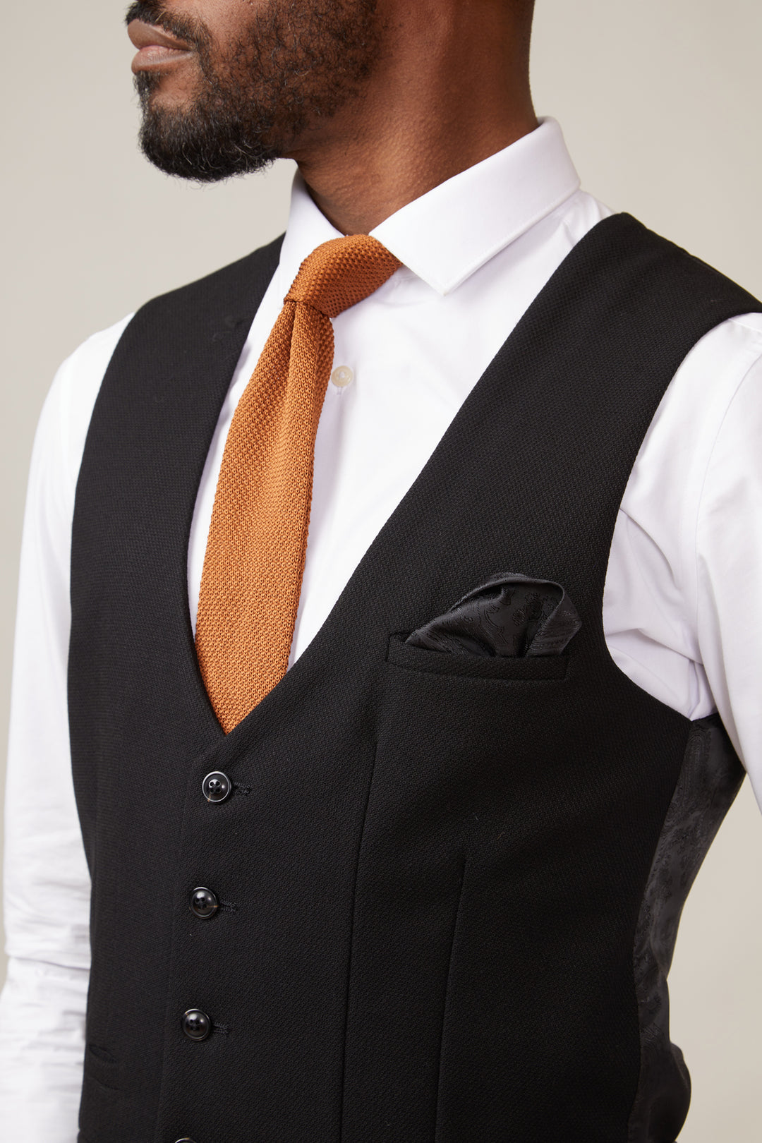 Man wearing a black waistcoat, white shirt, and brown tie against a neutral background