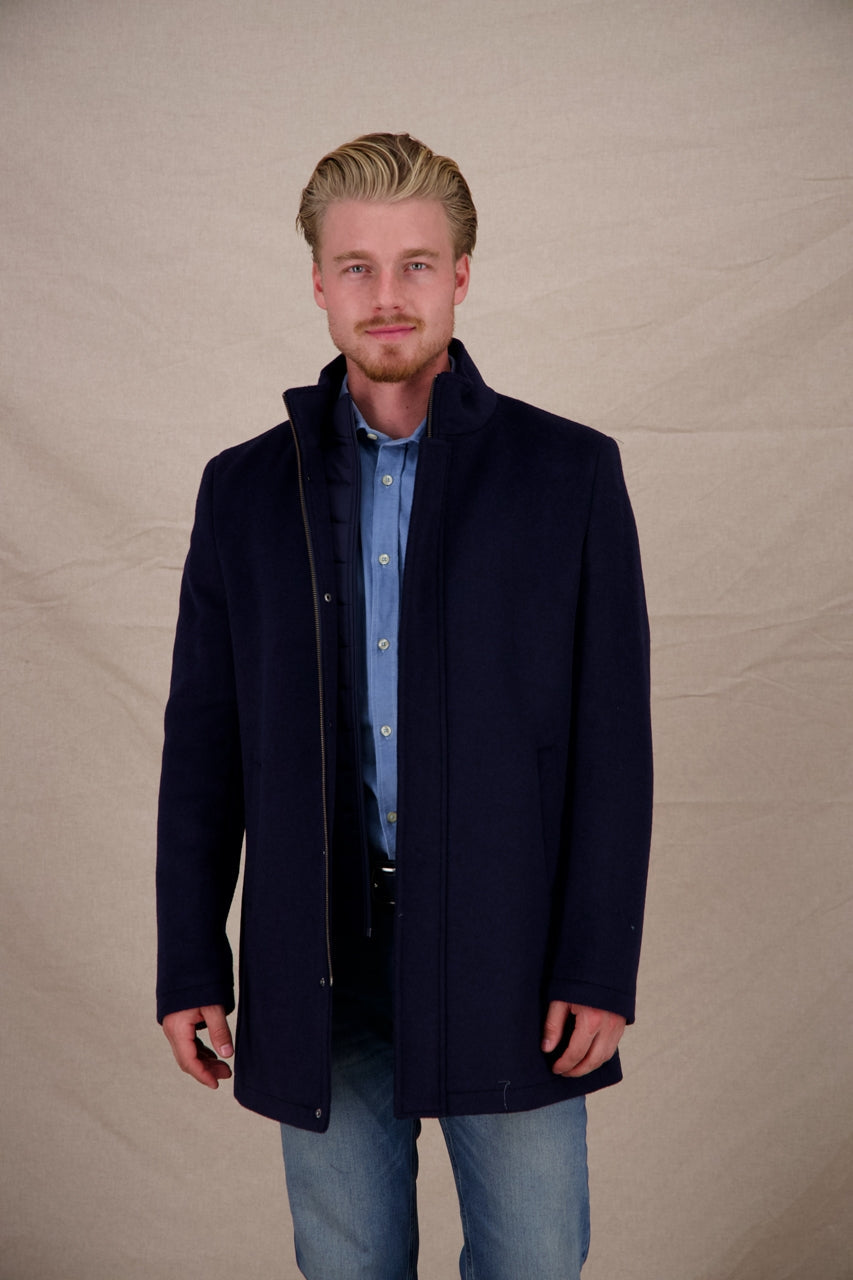 Man wearing a dark blue coat over a light blue shirt and jeans on a beige background