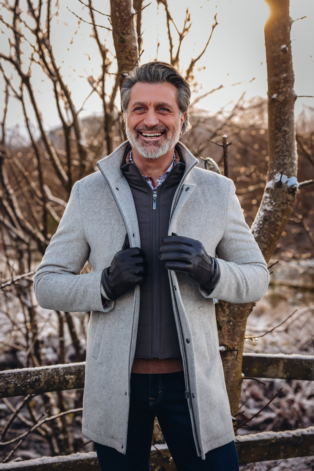 Man wearing a gray coat and gloves standing in a snowy landscape with bare trees.