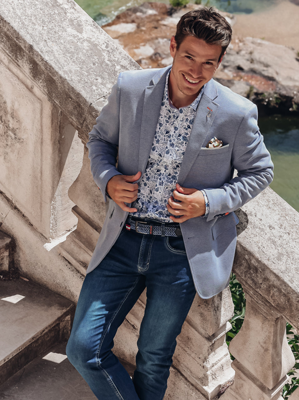 Man in a jeans blue blazer and blue jeans standing on a stone balcony.