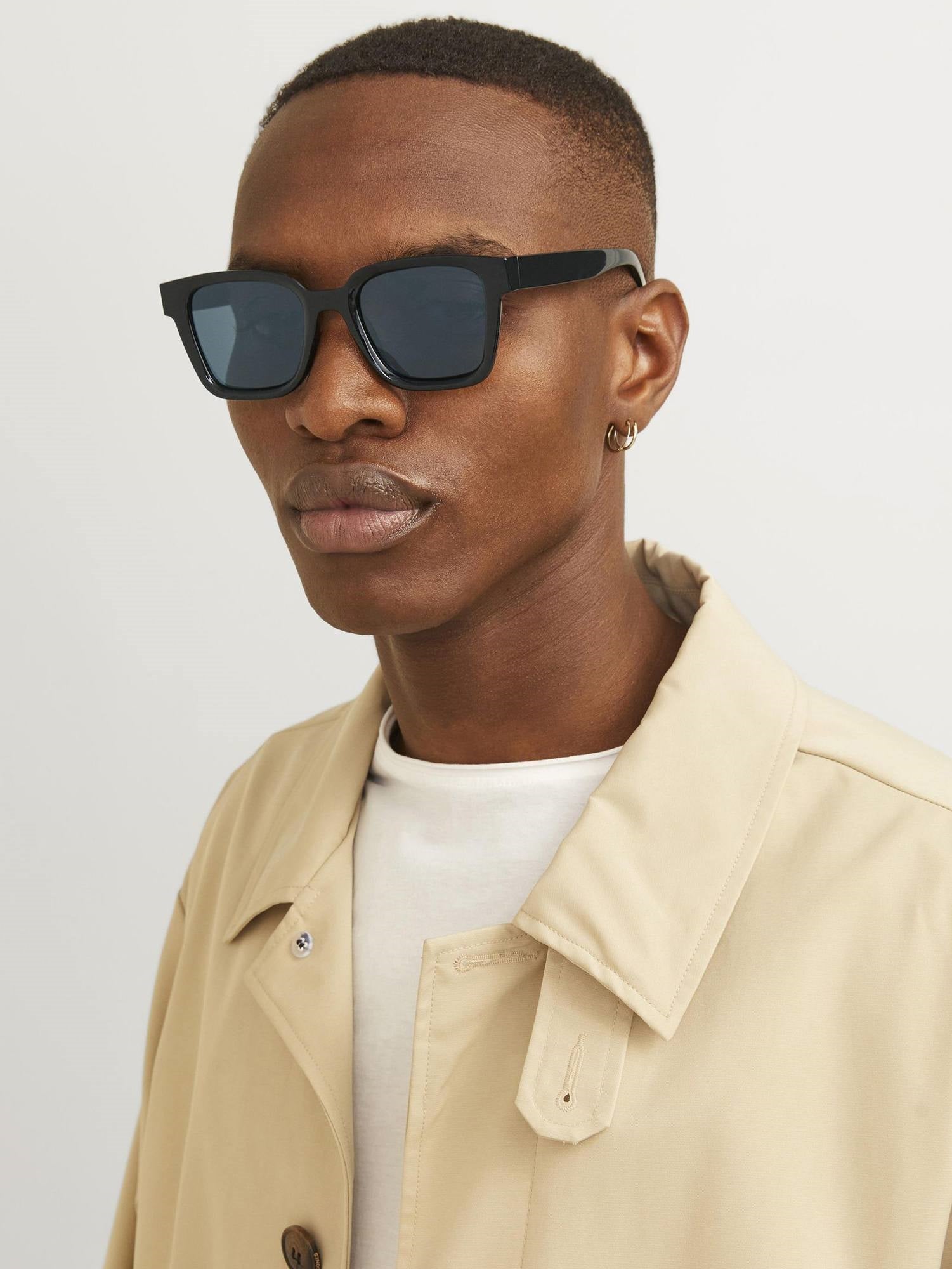Person wearing black sunglasses and a beige jacket on a plain background