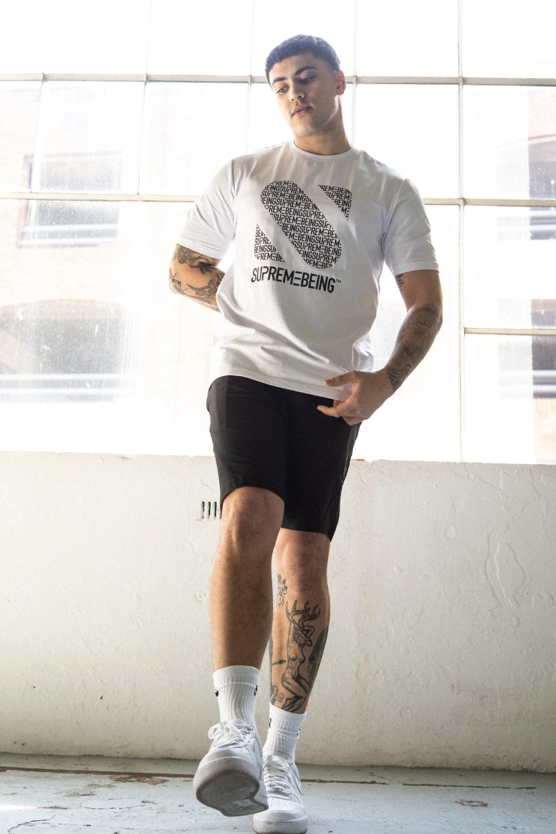 Person wearing a white t-shirt with a graphic design and text, black shorts, and white sneakers in a room with large windows.