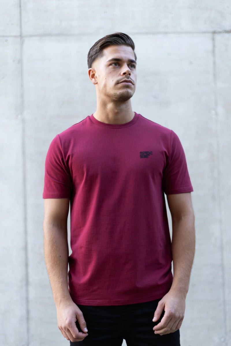 Man wearing a maroon t-shirt with a small logo against a gray concrete wall