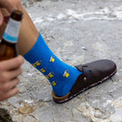 A person wearing a bright blue crew sock with a repeating pattern of yellow beer glasses and white foam, paired with a dark brown clog shoe, standing on a gray stone surface while holding a brown glass bottle.