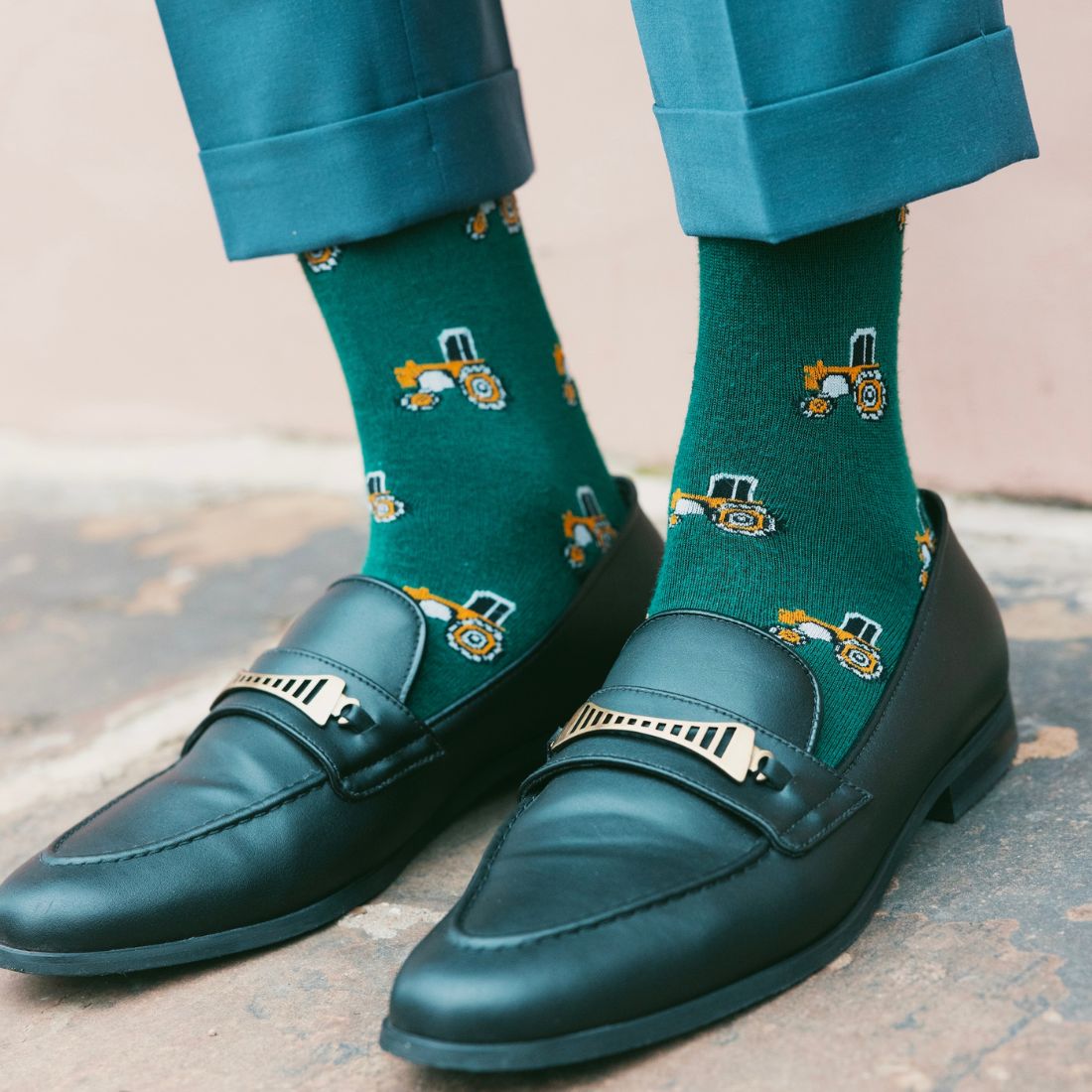 Teal loafers with decorative buckles worn with green socks featuring tractor designs on a textured surface.