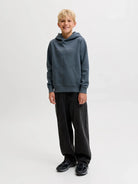 A young boy stands against a light gray studio background wearing a slate blue-gray pullover hoodie with a small white chest logo, black sweatpants, and black shoes.