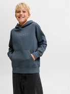 A smiling young boy with blonde hair wearing a slate blue-gray pullover hoodie with a small white dotted logo on the chest, paired with dark gray jeans, against a plain light gray studio background.