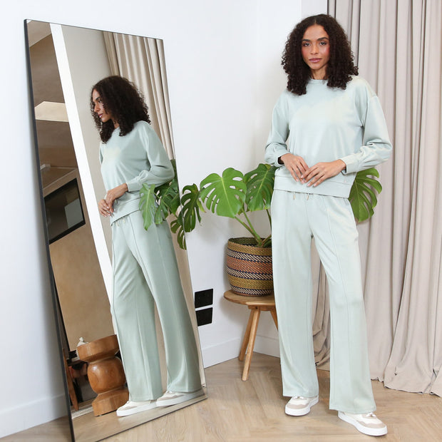Woman in a light green outfit standing next to a full-length mirror in a room with plants and decor.